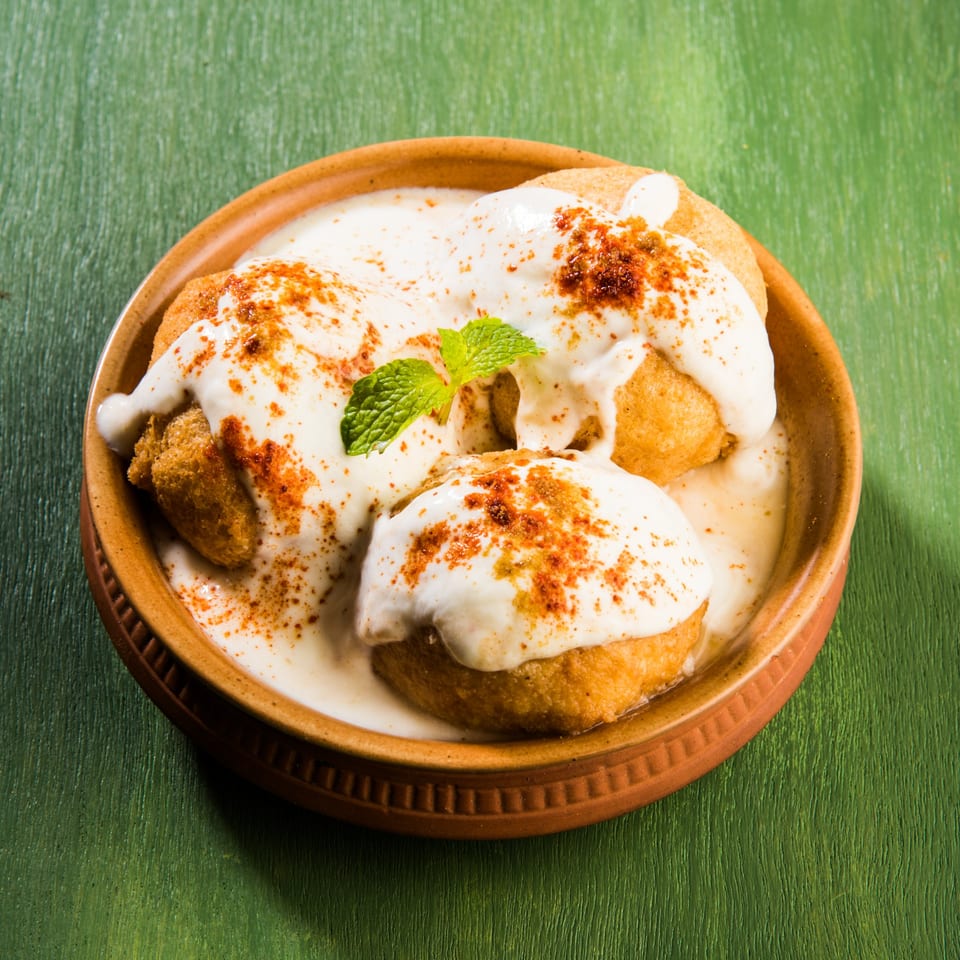 Dahi Vada, an Indian snack, with yogurt, spices, and mint in a brown bowl on a green surface.
